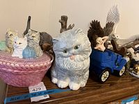 Photo showing assortment of animal figurines including cats in basket, large white Persian cat figurine, owl, eagle, and vintage blue toy car with figurines.