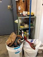 Photo showing two buckets containing multiple used baseball mitts, a blue handled slingshot, and part of the metal cart with assorted items on shelves.