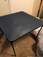 33 inch square black folding card table with scratches and stains on top surface.