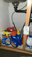 Photo under sink showing assorted cleaning bottles including OxiClean, Clorox, Tide, and a large white liquid bottle.