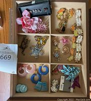 Top view of divided box with assorted costume jewelry including multiple brooches, earrings, and bracelets in various colors and styles.