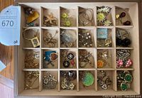 Overhead view of a compartmentalized box showing a variety of costume jewelry items including necklaces, earrings, brooches, and pendants in gold and silver tones with some colored accents such as green and pink.