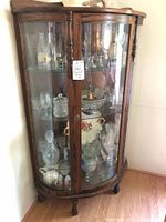 Full front view of vintage curved glass china cabinet showing glass display shelves with decorative glassware inside. Wooden frame with curved glass door and brass lock.