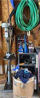 Packard drill press on stand with shelf behind holding funnels and auto supplies, green hose coil above