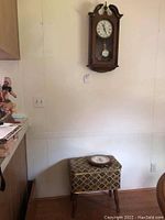 Howard Miller Westminster Chime wall clock mounted on wall above upholstered storage ottoman with a clock face and pendulum visible.