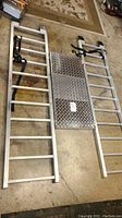 Full view of the landing ramp laid on the ground showing two aluminum ramp sections with a diamond plate platform in the middle, and black securing straps attached.