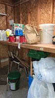 Wide shot showing boxed car oil containers, chemical bottles, paper bags, and white plastic bucket on a wooden shelf in a plywood-walled room.