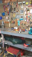 Wide shot of pegboard with assorted tools including pliers, screwdrivers, paint scrapers, and funnels hanging, workbench with items, green tools case below