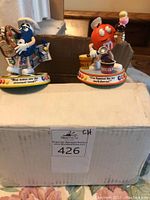 Two M&M character figurines: Blue M&M playing saxophone and Red M&M holding spoons with vanilla and chocolate containers on bases with phrases.