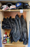 Open metal storage cabinet with two black leather jackets hanging, folded leather chaps and pants on shelf, assorted maintenance supplies on bottom shelf.