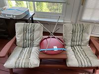 Overall photo showing the full brown wooden patio bench with two seat and two back removable outdoor fabric cushions with vertical stripes and button tufting. The matching brown wooden side table sits between the chairs with a red round metal tray strapped upside down on top.