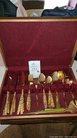 Photo showing full set of gold colored silverware in red velvet lined case with knives, forks, spoons, ladle and tongs.