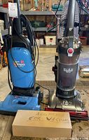 Two upright Hoover vacuum cleaners side by side in a workshop or storage space with a box of vacuum attachments in front labeled for vacuum.