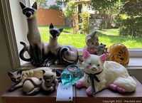 Full view of vintage ceramic cat figurines and decor arranged on windowsill with outdoor background showing the Siamese cat lamp and various other cat figurines