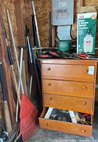 Wooden chest with three drawers, assorted long-handled yard tools including leaf rake, shovels, and rakes leaning against wall.