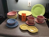 Shows a stack of colorful 10.5-inch Fiesta dinner plates including orange, yellow, green, and purple, with pink and yellow berry bowls, blue cereal bowls, and two pink cups with saucers.
