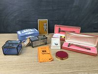 Main overview photo showing multiple items including leaded glass boxes, Jasperware-style box, little devil pendant, small white box with earrings, round red compact, and two boxed razors.