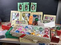 Photo of multiple boxed glass Christmas ball ornaments showing various colors, packaging styles, and designs including glitter and metallic finishes.