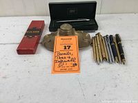 Full lot on table with box of pencils on left, brass inkwell center, pens arranged to right, green Cross pen box in background.