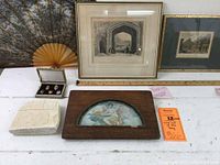 Overall lot arrangement showing framed prints, Asian fan, enameled lockets in box, and framed art on table with ruler for scale.