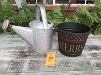 Photo showing vintage Dover watering can and painted herb bucket placed on rustic wooden surface outdoors with greenery background.