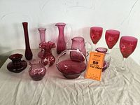 Full view of all 12 red glass pieces on white cloth background, showing various shapes and styles including vases, pitchers, basket and wine glasses.