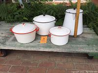 Four white enamelware kitchen items on a rustic bench: two pots with red accents, one pot with black accent, and one tall coffee pot with black trim.