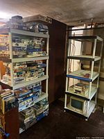 Two white plastic shelving units positioned side by side in a basement; one fully loaded with various boxed items, the other mostly empty with a computer monitor on the bottom shelf.