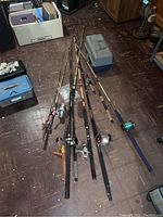 Multiple fishing rods laid on the floor, some with reels attached, alongside a blue and gray plastic tackle box.