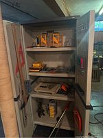 Overall view of open metal storage cabinet containing tools and hardware on shelves and inside door racks.