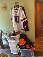 Two college jerseys hanging on black hangers with a white jersey in front marked number 10, maroon and gold colors, size XL by Colosseum Athletics, with other jersey behind. Below are plastic storage bins and baskets containing multiple hats including a bright orange hat.