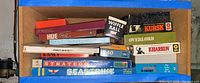 Top view of cardboard box containing assorted board games including Stratego, military campaign games like Hue, Operation Pegasus, Kursk, Overlord, Kharkov, Scrabble and Wiffle Ball set.