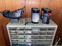 Overall view of black miniature ship, model spray paints, and plastic storage drawers against wooden wall
