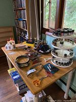 Wide view of wooden table displaying paints, stands, rulers, pliers, scissors, and adhesives with window background