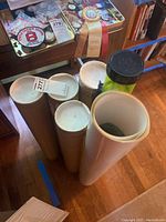 Photo showing several poster tubes in various colors and sizes standing upright on a wooden floor near a table.