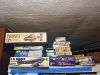 Photo showing various scale model kits stacked including the Tamiya Tiger I tank kit box, Monogram C-47 Skytrain and Paratroopers, and Lindberg U.S.S Olympia kits