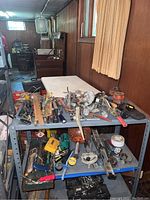 Wide view showing tables with assorted used hand tools including wrenches, clamps, hammers, saws and screwdrivers placed on metal shelving and table.