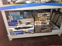Photo of stacked boxed model kits including World War II aircraft and military vehicles under a plastic shelf.