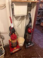 Photo showing two upright vacuum cleaners: a red Hoover WindTunnel on the right and the Hoover Elite Rewind on the left with visible attachments and rewind cord feature.