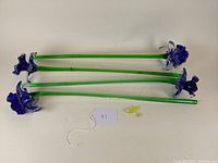 Four long green glass stems with blue glass flower heads and scattered loose yellow glass stamens on white surface.