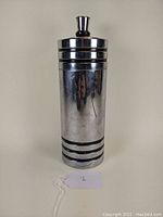 Full frontal view of the tall chrome cocktail shaker showing its cylindrical form and horizontal groove details