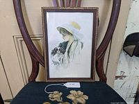 Framed vintage print showing a woman in a large hat holding a spaniel dog, displayed on a dark cushioned chair with wooden backrest.