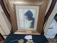 Framed hand tinted photograph showing full view of the frame and image of woman looking out window, set on a chair.