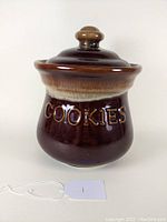 Brown ceramic cookie jar with lid, displaying "COOKIES" embossed on the side in raised lettering and visible drip glaze on top rim.