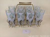 Eight highball tumor glasses arranged in the brass wire caddy, showing foliage leaf decoration on glasses and holder design.