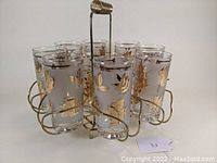 View of eight clear glass tumblers with gold leaf and frosted etched foliage pattern arranged in brass wire caddy