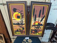 Pair of 1970s framed wall art prints showing stylized sunset scenes with silhouetted plants on striped backgrounds. One print has cattails, the other has wildflowers/thistle.