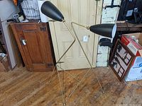 Pair of Mid-Century Modern style floor lamps with thin brass legs and black cone shades standing on a wooden floor against a cream-colored door.