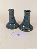 Two blue glazed studio pottery candlesticks with ringed body detail, slightly different shapes, placed on a pale surface with a tag numbered 95 visible