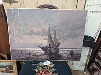 Full view of oil painting showing two sailboats at shore under cloudy sky, resting on a green surface with other artworks around.